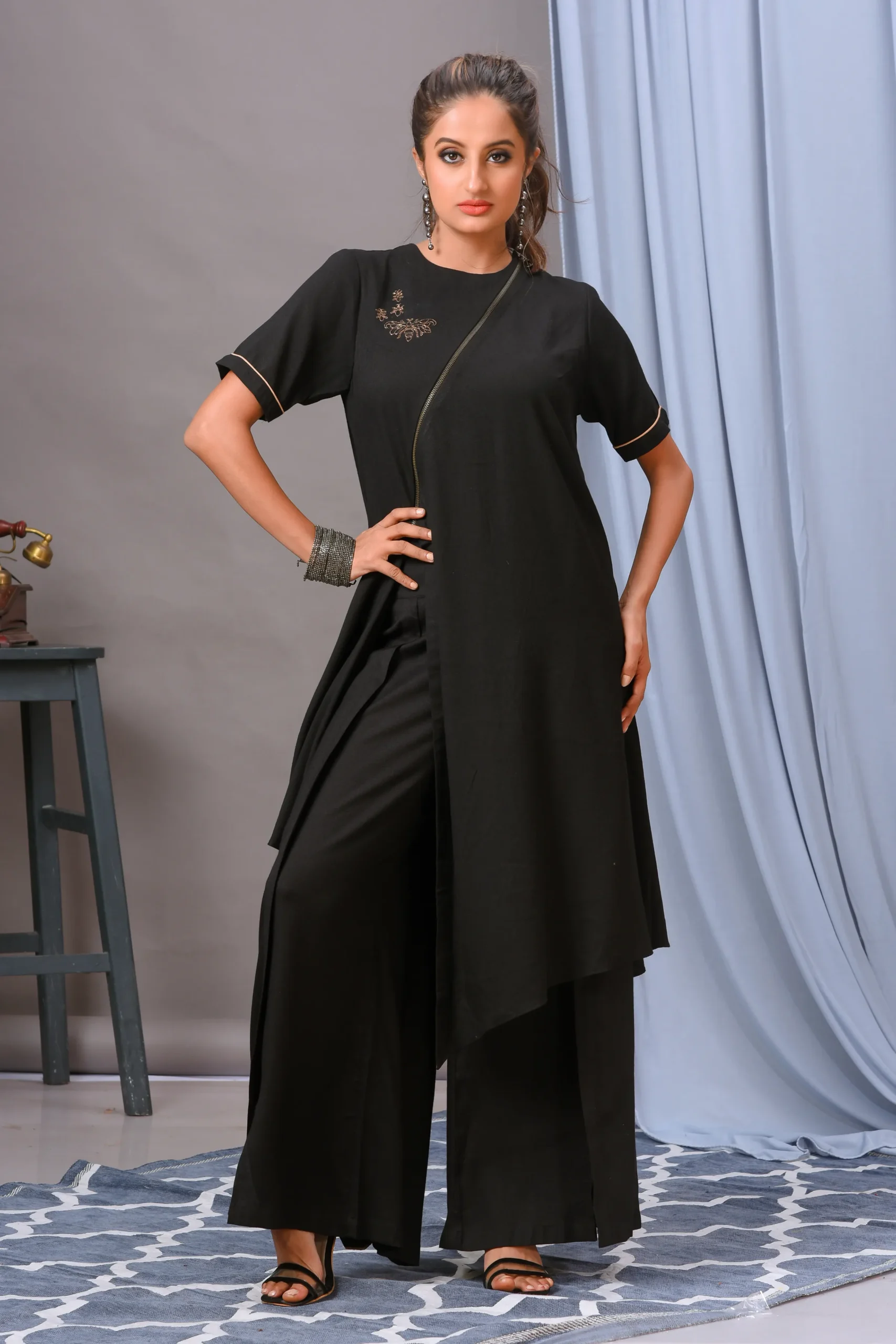 A black cotton rayon front-open asymmetric tunic from Golden Muslin By Vivek Kumar, featuring minimal hand embellishments and paired with chic black pants.