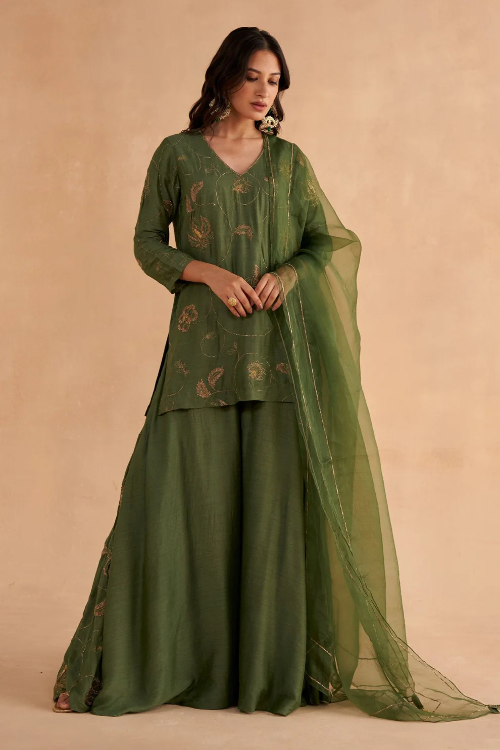 “Golden Muslin fine Moonga silk kurta with all-over Zardozi chintz-inspired embroidery, paired with bottle-green Moonga silk Sharara pants with waist belt, pockets, slit, and side Zardozi work, completed with bottle-green organza dupatta with golden border and embroidered tassels.”