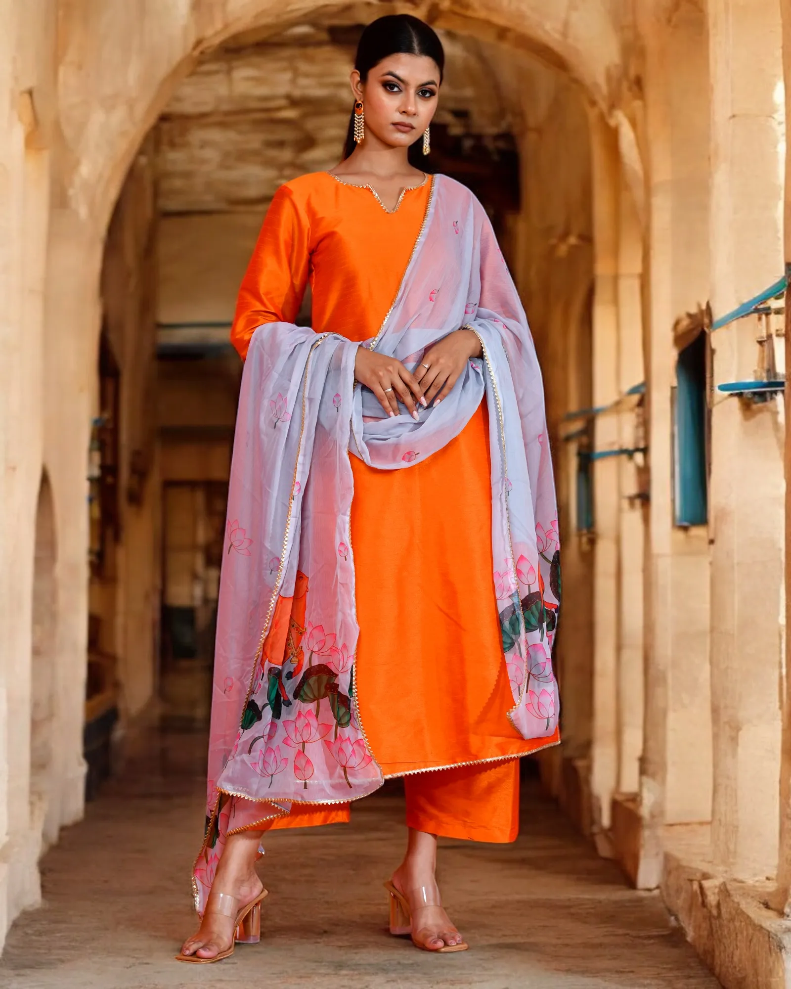 A structured orange dupion suit set paired with a grey Pichwai printed chiffon dupatta from Golden Muslin by Priyanka Rathore, blending heritage artistry with festive elegance.
